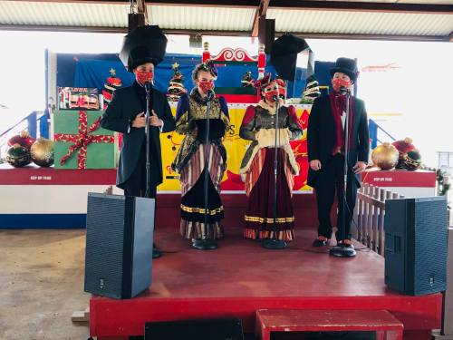 Four performers in festive attire sing on stage, surrounded by holiday decorations and speakers.