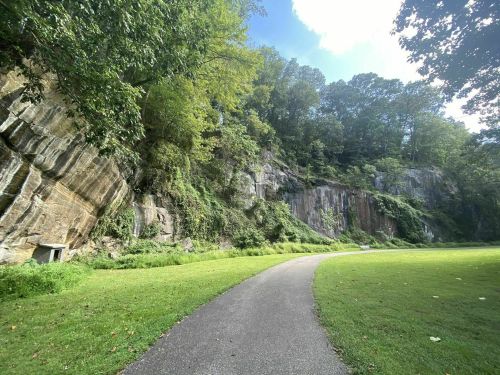 A winding path through a lush green park, flanked by rocky cliffs and trees under a bright blue sky.