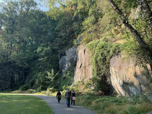 Three people walk along a path beside a rocky cliff, surrounded by trees in a sunny park setting.