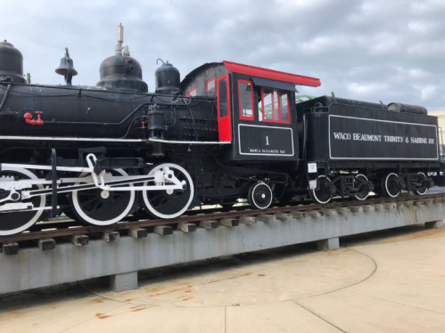 A vintage black steam locomotive with a red cab, displayed on a platform against a cloudy sky.