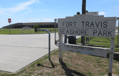Sign for Fort Travis Seashore Park with a stop sign and grassy area in the background under a clear blue sky.