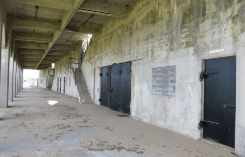 Abandoned concrete structure with black doors and a staircase, featuring a sandy floor and weathered walls.