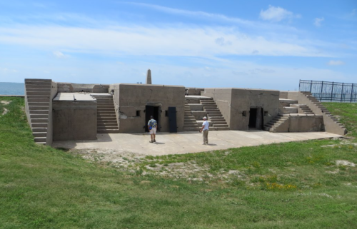 Two people stand near a concrete structure with stairs, overlooking a grassy area and a body of water under a blue sky.