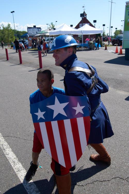 A man in a superhero costume poses with a boy holding a shield, both smiling at an outdoor event.