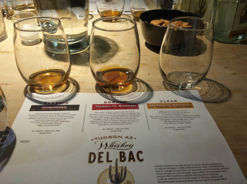 Three whiskey glasses with different shades of amber liquid, alongside a tasting menu for Del Bac whiskey.