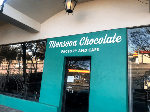 Exterior of Monsoon Chocolate Factory and Cafe, featuring a teal wall and large white sign. Windows reflect the surroundings.