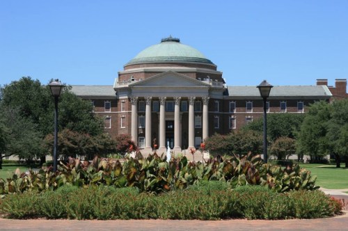 A large brick building with a dome, surrounded by greenery and flowers, under a clear blue sky.