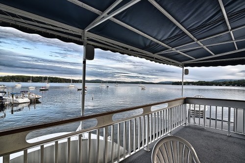 View from a covered deck overlooking a calm lake with boats and distant hills under a cloudy sky.