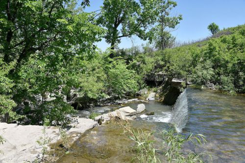 A serene landscape featuring a small waterfall surrounded by lush greenery and clear water under a blue sky.