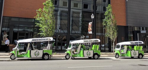 Three green and white tuk-tuks parked along a city street, with trees and a modern building in the background.