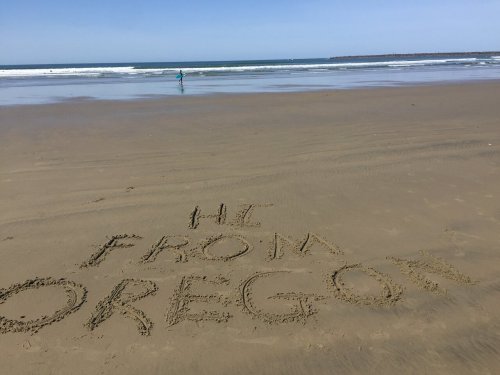 Text written in the sand on a beach reads "HI FROM OREGON," with a person walking in the background near the water.