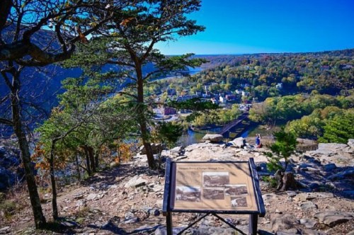 Scenic view from a rocky overlook, featuring a historical sign and a vibrant landscape of trees and a river below.