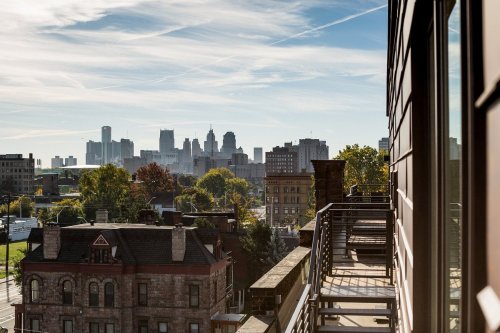 View of a city skyline with modern and historic buildings, framed by trees and a balcony railing in the foreground.