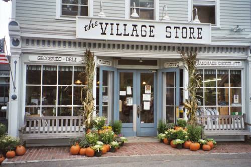 A quaint village store with a welcoming entrance, decorated with pumpkins and flowers, showcasing groceries and handmade items.