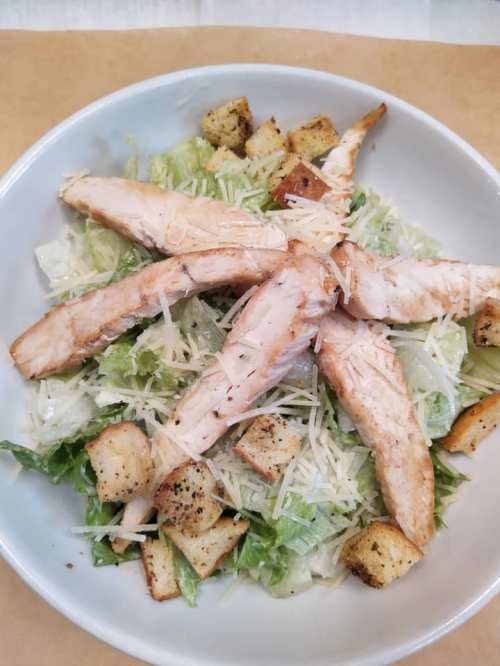A bowl of Caesar salad topped with grilled chicken strips, croutons, and grated Parmesan cheese on a bed of lettuce.