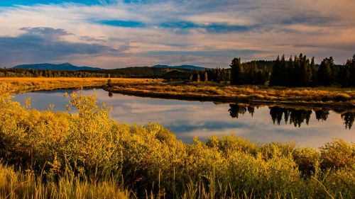 A serene river reflects golden foliage and mountains under a blue sky with scattered clouds.