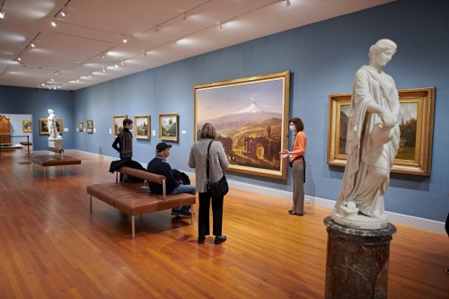 Visitors admire artwork in a gallery, with a statue in the foreground and paintings displayed on the walls.