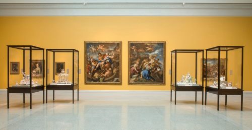 Art gallery interior featuring paintings on yellow walls and display cases with sculptures and decorative art.