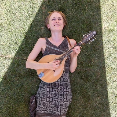 A woman lies on grass, playing a mandolin and smiling, wearing a patterned dress and sunglasses.