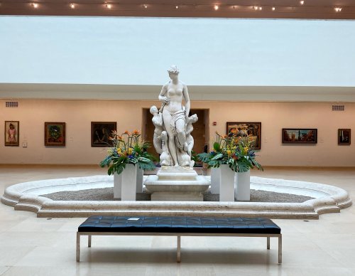 A white marble statue of a woman stands in a gallery, surrounded by vibrant flowers and paintings on the walls.