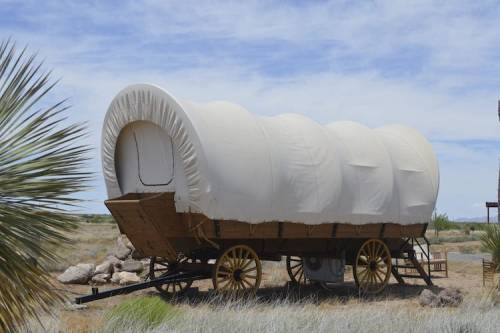 A covered wagon on wheels, set in a grassy area with sparse vegetation and a blue sky in the background.