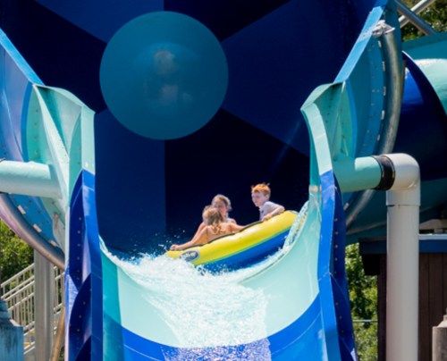 Three children slide down a colorful water slide, splashing into the water below on a sunny day.
