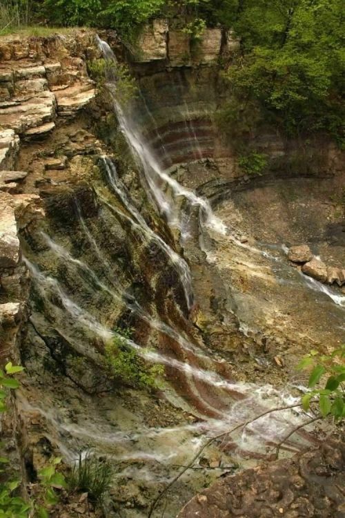 A cascading waterfall flows over rocky cliffs, surrounded by lush greenery and a serene natural landscape.