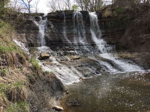 A serene waterfall cascading over rocky ledges into a calm pool, surrounded by greenery and trees.