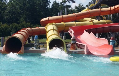 Colorful water slides in a pool, with splashes of water and greenery in the background on a sunny day.