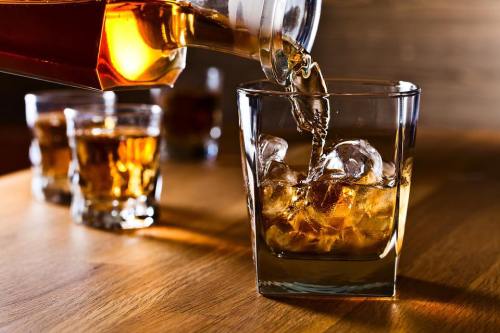 A glass being filled with whiskey over ice, with additional glasses of whiskey in the background on a wooden table.
