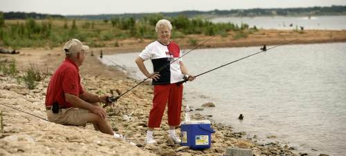 An elderly woman and man fish by a lake, with the woman standing and the man sitting on the shore.