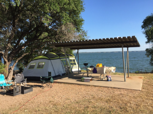 A campsite by a lake featuring a tent, picnic table, and shaded area with chairs under a clear blue sky.