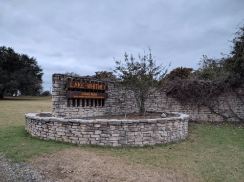 Sign for Lake Whitney State Park, featuring stone structure and surrounding greenery under a cloudy sky.