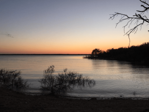 A serene lake at sunset, with calm waters reflecting soft hues and silhouettes of trees along the shore.