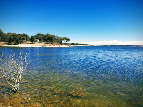 A serene lake scene with clear blue water, rocky shore, and trees under a bright blue sky.