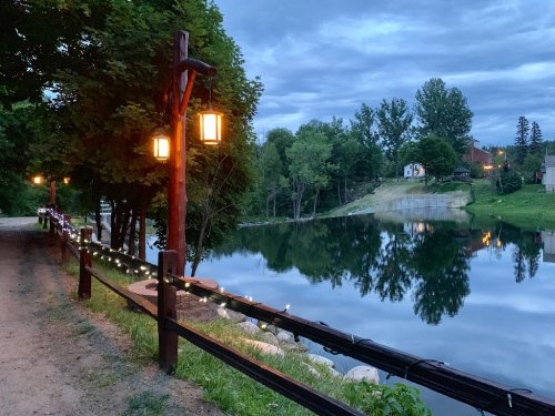A serene lakeside path at dusk, lined with lanterns and fairy lights, reflecting trees and clouds in the calm water.