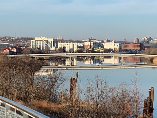 A calm river reflects buildings and a bridge under a clear sky, surrounded by bare trees and urban scenery.