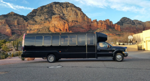 A long black bus parked on a road with red rock mountains in the background under a blue sky.