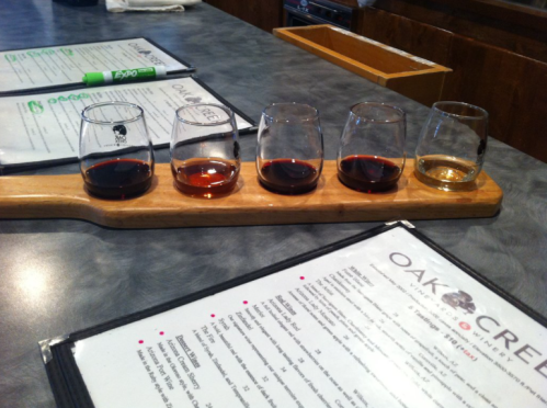 A wooden flight board with five small glasses of different colored liquids, placed on a table with menus.