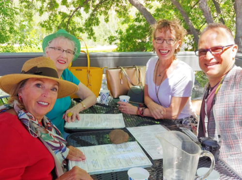Four friends sitting at an outdoor table, smiling and looking at menus, surrounded by greenery.