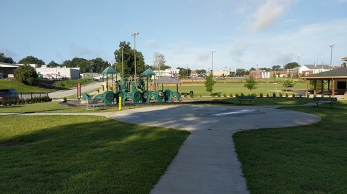 A playground with green equipment in a park, surrounded by grass and a paved walkway, under a clear blue sky.