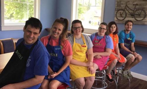 A group of six people in colorful aprons sitting on bar stools in a bright, cheerful cafe setting.