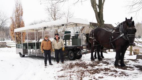 Two men stand beside a horse-drawn wagon in a snowy landscape, with trees and a wooden structure in the background.