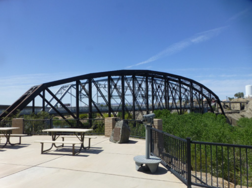 A black metal arch bridge spans over a river, surrounded by greenery and a paved area with picnic tables.