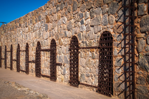 A stone wall with multiple arched windows, each secured with metal bars, under a clear blue sky.