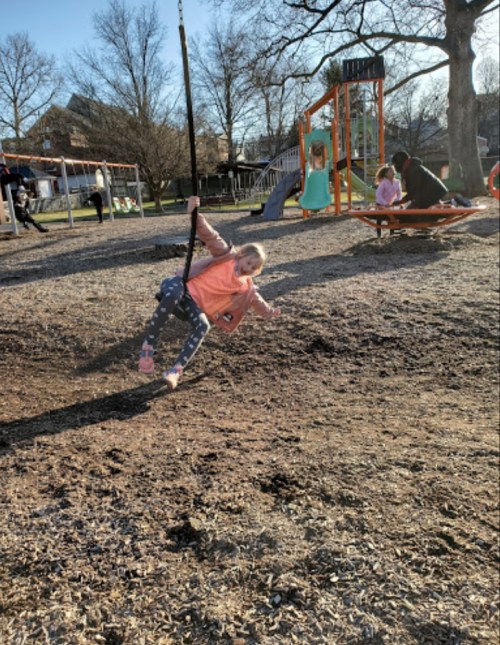 A child swings joyfully at a playground, with other children and play structures in the background on a sunny day.