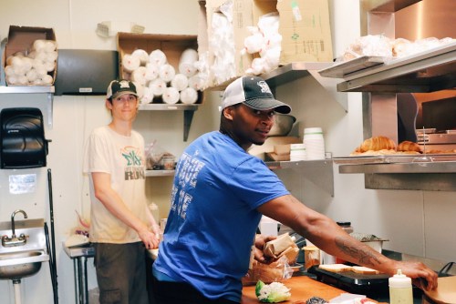 Two men working in a kitchen, one preparing food and the other standing nearby, with kitchen supplies in the background.