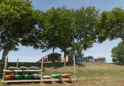 A row of colorful cabins behind a grassy area with kayaks stacked on a wooden rack under large trees.