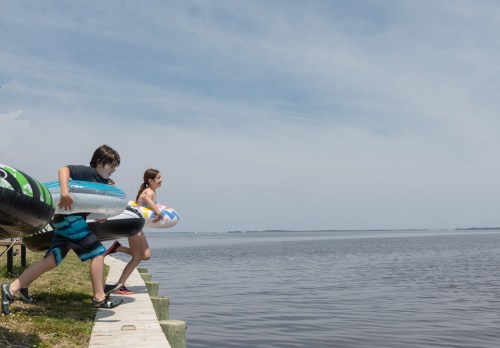 Two children run towards a calm lake, carrying inflatable floaties on a sunny day.