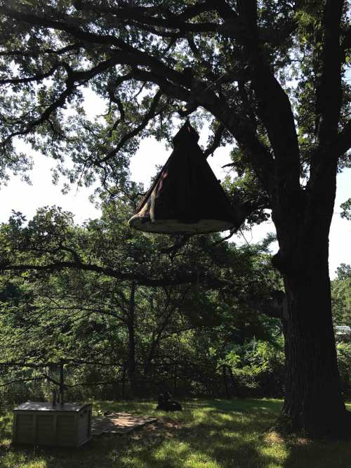A unique treehouse shaped like a tent hangs from a large oak tree, surrounded by greenery and sunlight.